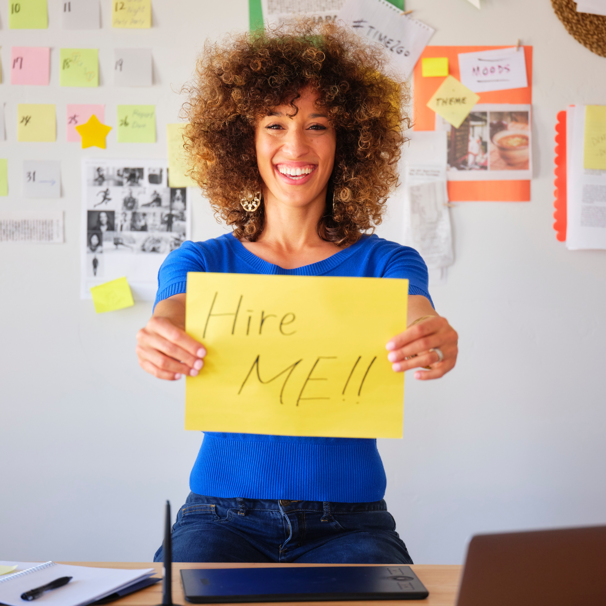 Image of a smiling woman in front of a wall holding a yellow sign that says "Hire Me!!" for the blog "5 Diversity Hiring Mistakes to Avoid When Interviewing Diverse Applicants"